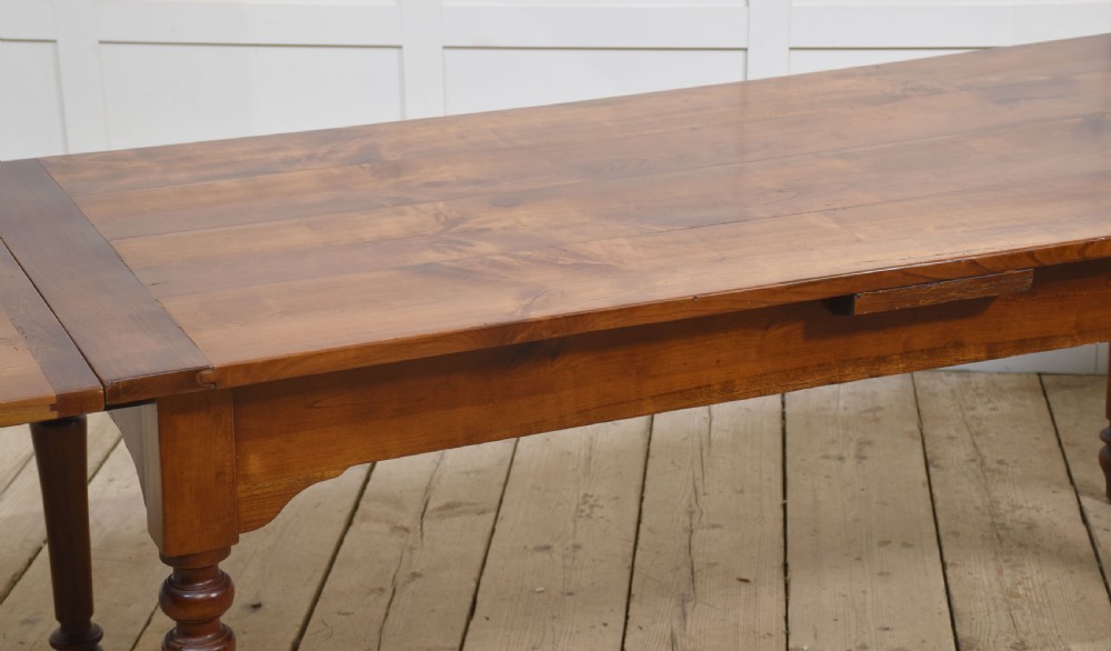 A large antique fruitwood farmhouse dining table featuring a smooth planked top, straight apron, and four turned legs resting on exposed wooden floorboards. View III