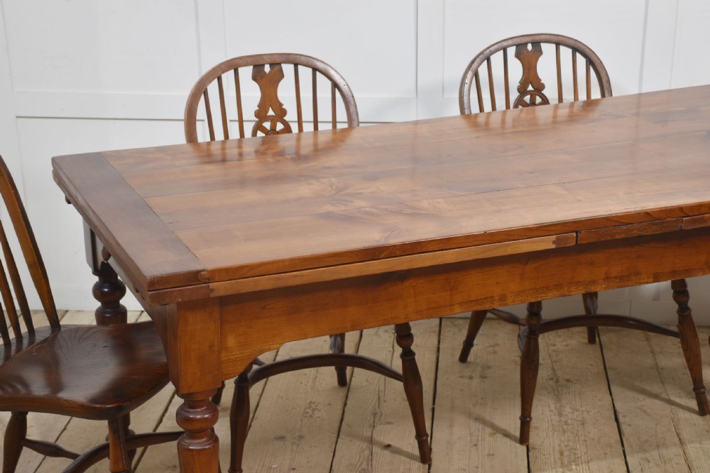 A large antique fruitwood farmhouse dining table featuring a smooth planked top, straight apron, and four turned legs resting on exposed wooden floorboards. View IV