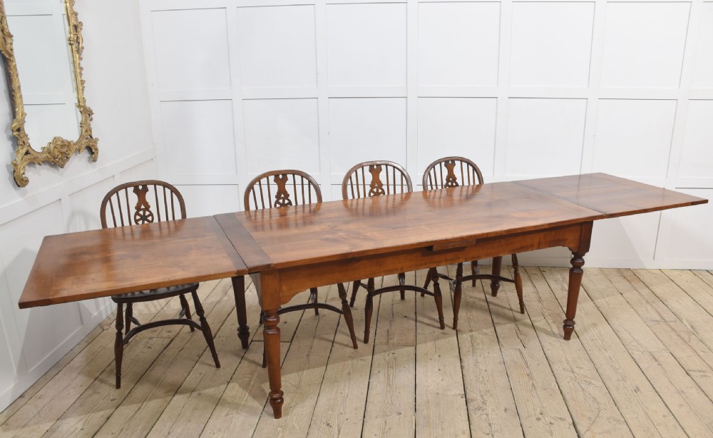 A large antique fruitwood farmhouse dining table featuring a smooth planked top, straight apron, and four turned legs resting on exposed wooden floorboards. View V