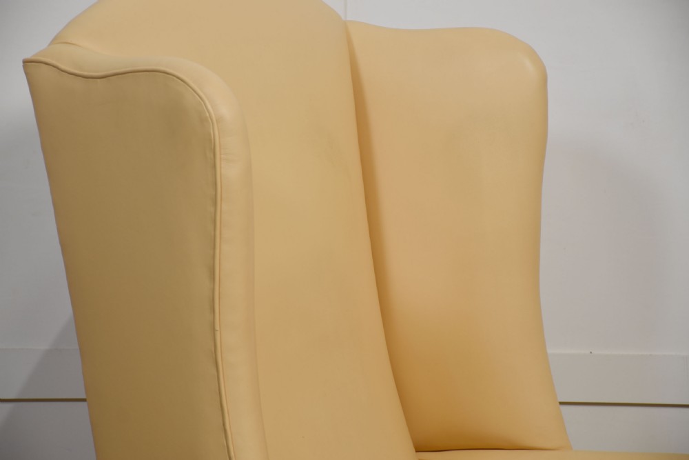 A high-backed cream leather wingback armchair with a fitted seat cushion and dark timber legs on brass castors. View VII