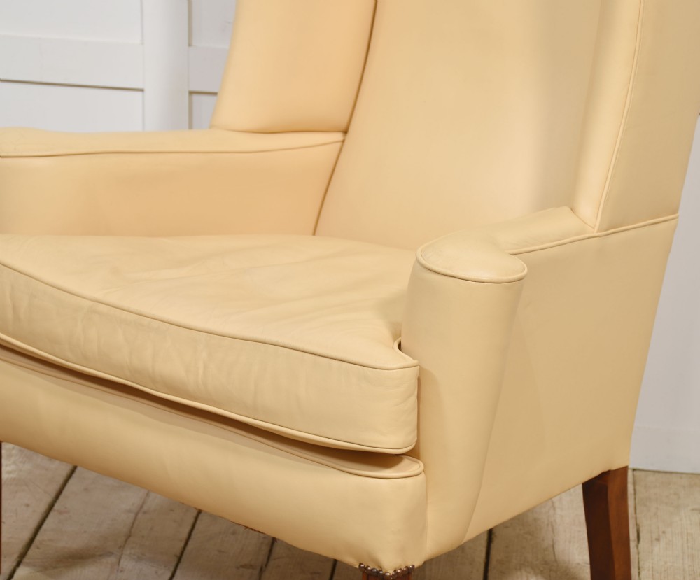 A high-backed cream leather wingback armchair with a fitted seat cushion and dark timber legs on brass castors. View X