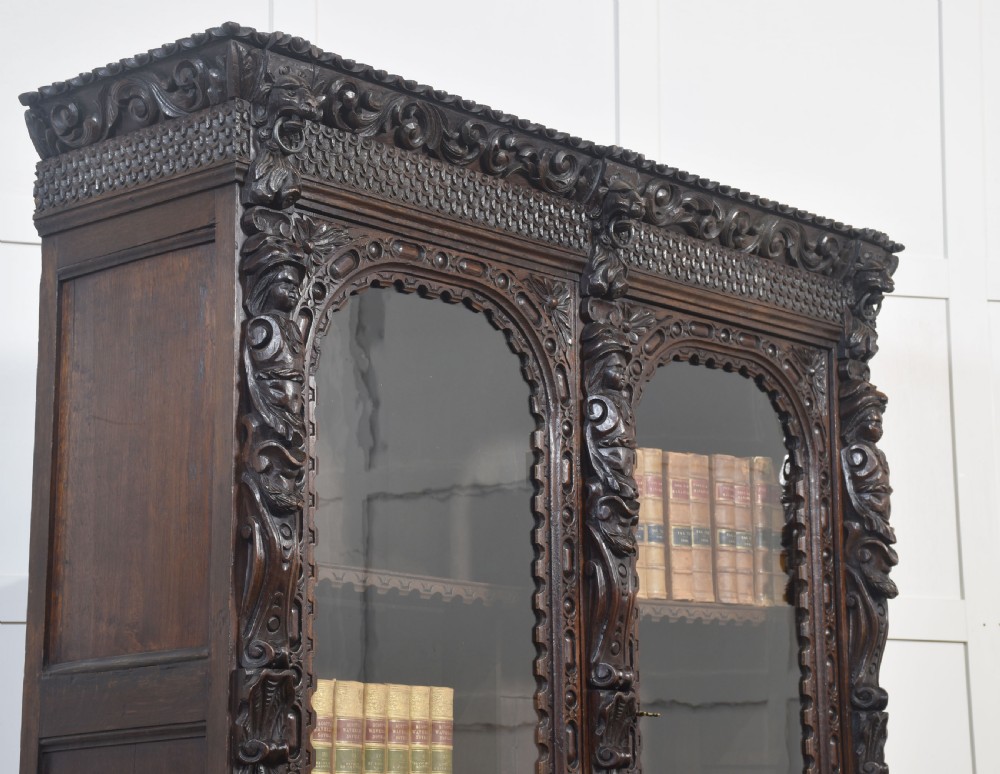 A tall dark oak Renaissance Revival bookcase featuring two glazed upper doors, intricate carved relief details, and geometric carved lower cupboard panels. View V
