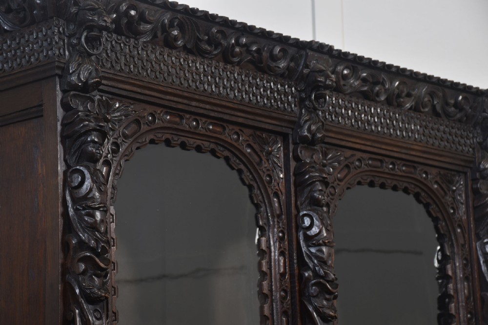 A tall dark oak Renaissance Revival bookcase featuring two glazed upper doors, intricate carved relief details, and geometric carved lower cupboard panels. View X