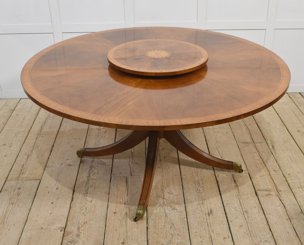 A circular mahogany dining table featuring crossbanded veneer edging, a central revolving lazy susan with marquetry inlay, and a pedestal support with four reeded legs terminating in brass castors.