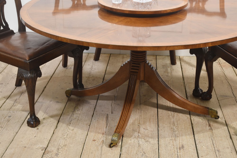 A circular mahogany dining table featuring crossbanded veneer edging, a central revolving lazy susan with marquetry inlay, and a pedestal support with four reeded legs terminating in brass castors. View X