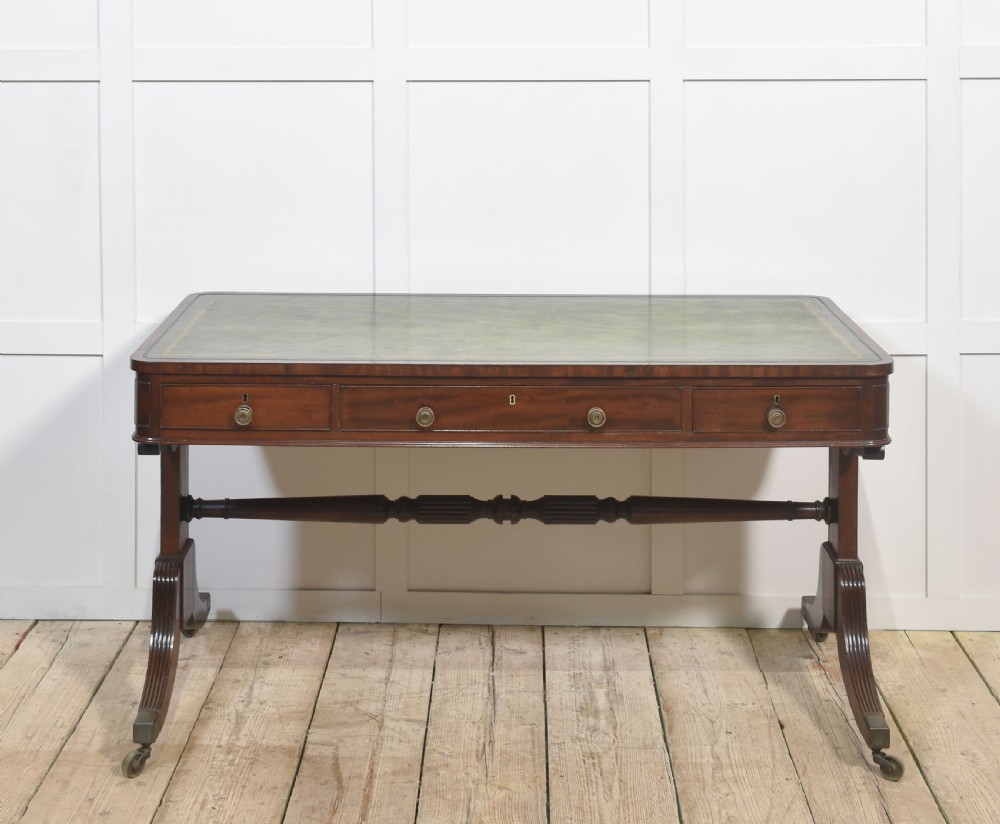 A Regency mahogany library table featuring a green leather inset top, brass knob drawers, a turned central stretcher, and reeded sabre legs on brass castors.