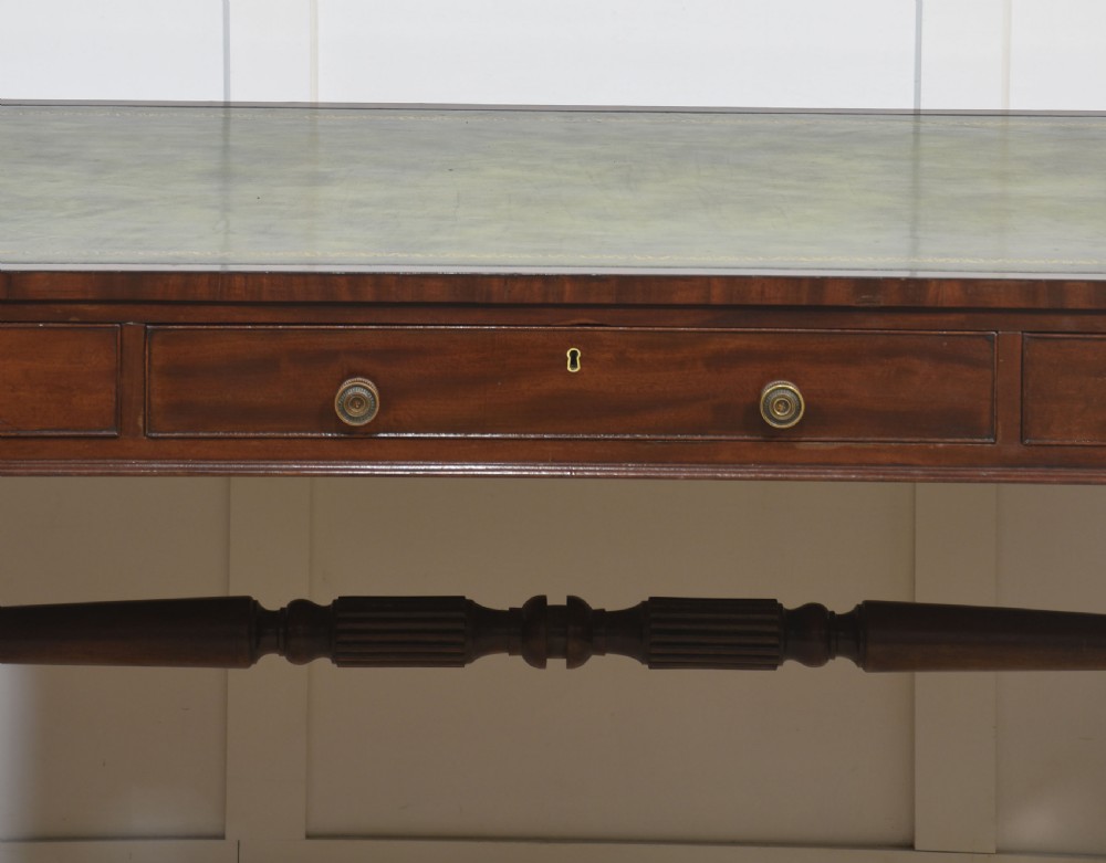 A Regency mahogany library table featuring a green leather inset top, brass knob drawers, a turned central stretcher, and reeded sabre legs on brass castors. View VII