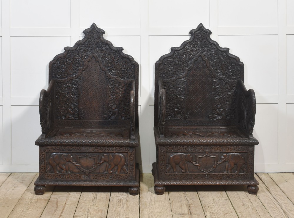 A pair of 19th-century dark hardwood ceremonial armchairs featuring intricate relief carvings, arched backrests, and elephant motifs on the base panels.