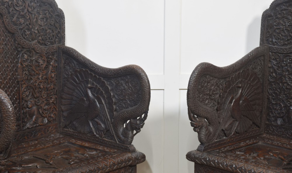 A pair of 19th-century dark hardwood ceremonial armchairs featuring intricate relief carvings, arched backrests, and elephant motifs on the base panels. View X