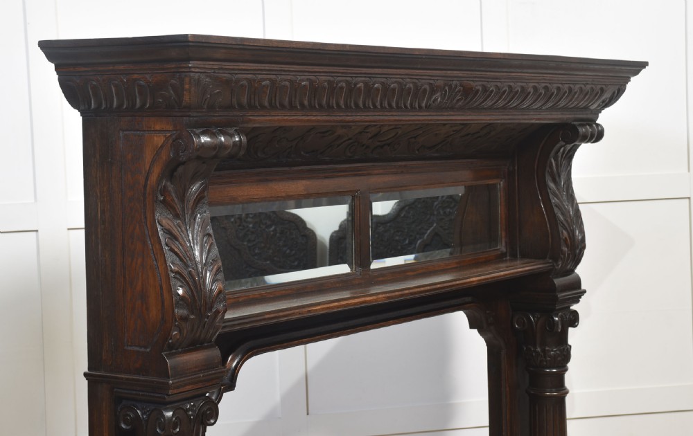 A carved dark oak fire surround featuring twin mirror panels set above a wide hearth opening, framed by fluted columns and detailed leaf carvings. View XVI