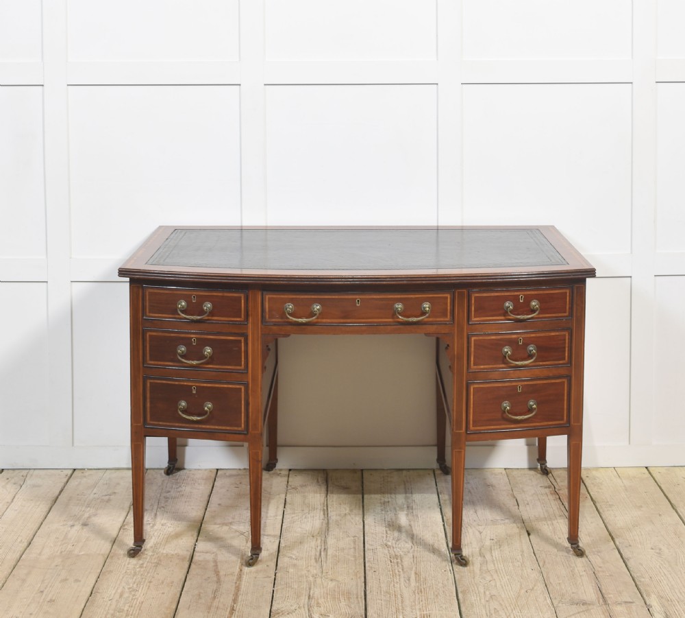 An antique mahogany writing desk featuring a leather insert top, boxwood stringing inlay, seven drawers with brass handles, and tapered legs on castors.