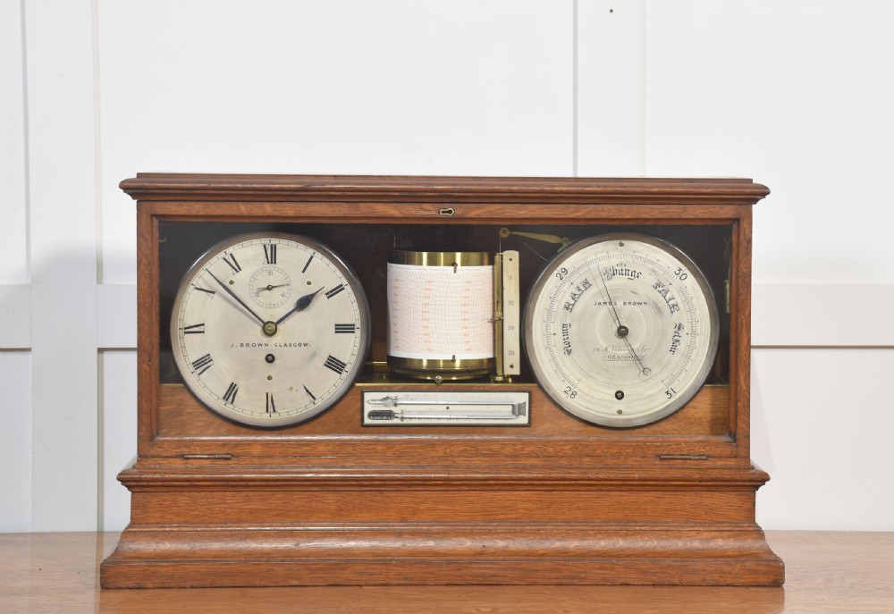 A glazed oak-cased antique weather station featuring a clock dial on the left, an aneroid barometer on the right, and a central paper drum barograph with an integrated thermometer.
