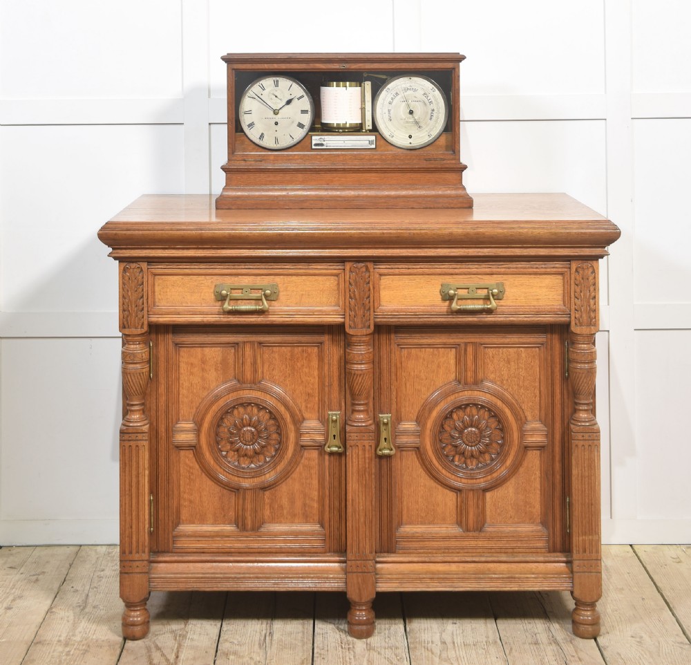 A glazed oak-cased antique weather station featuring a clock dial on the left, an aneroid barometer on the right, and a central paper drum barograph with an integrated thermometer. View II