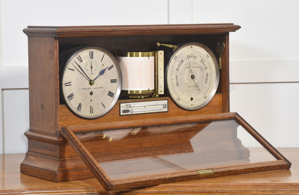 A glazed oak-cased antique weather station featuring a clock dial on the left, an aneroid barometer on the right, and a central paper drum barograph with an integrated thermometer. View VI