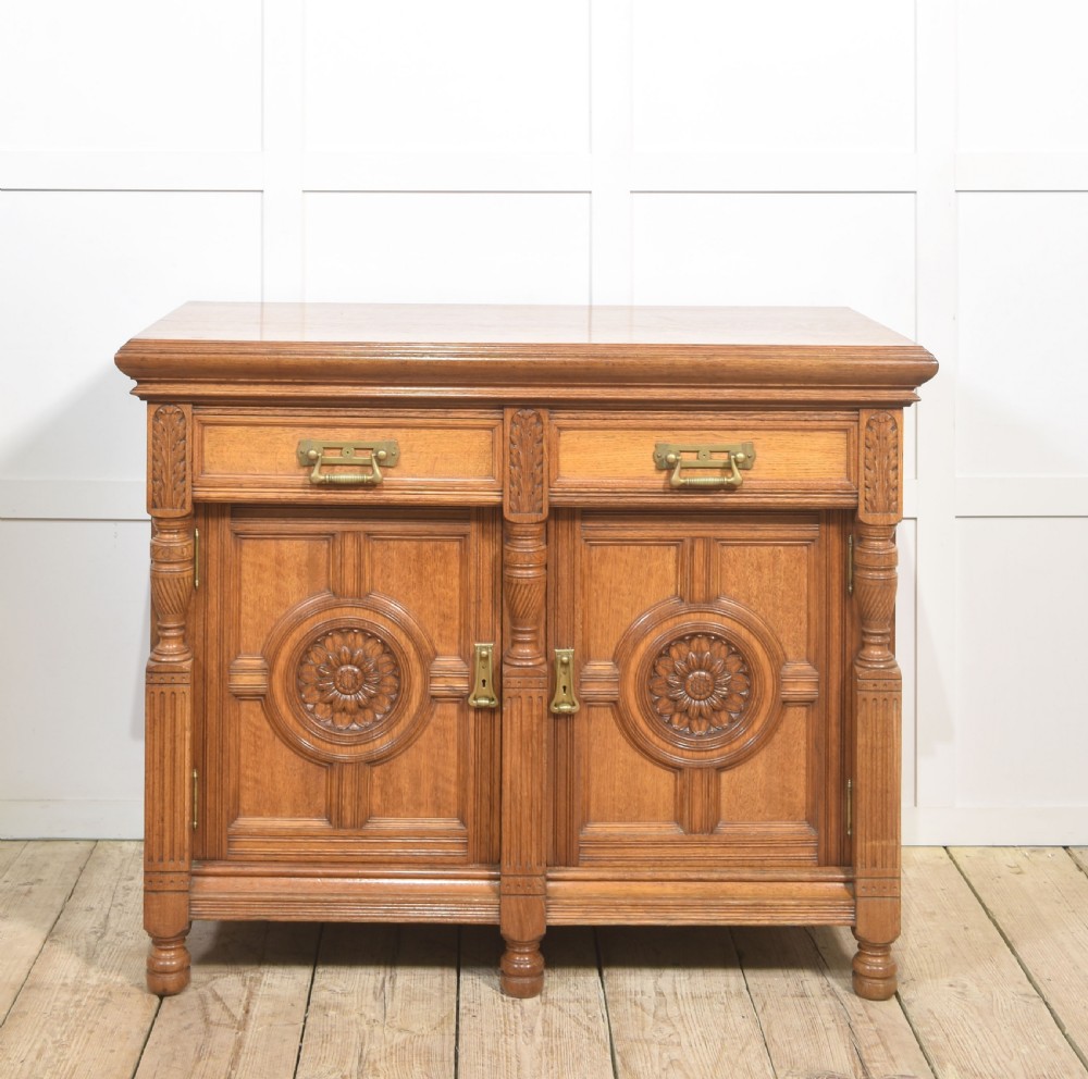 A carved oak two-door sideboard featuring two drawers with brass handles, central rosette carvings on the cupboard doors, and turned columns.