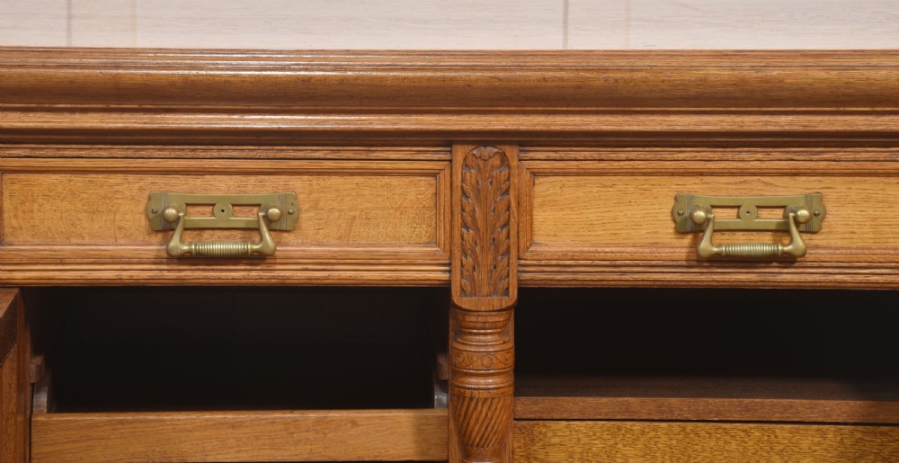 A carved oak two-door sideboard featuring two drawers with brass handles, central rosette carvings on the cupboard doors, and turned columns. View VI