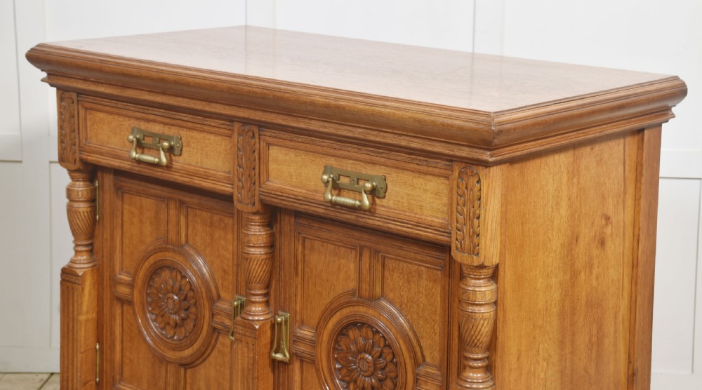 A carved oak two-door sideboard featuring two drawers with brass handles, central rosette carvings on the cupboard doors, and turned columns. View XVI