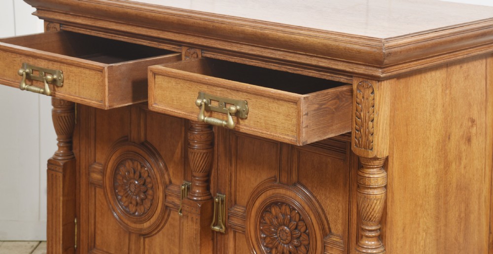 A carved oak two-door sideboard featuring two drawers with brass handles, central rosette carvings on the cupboard doors, and turned columns. View XVII