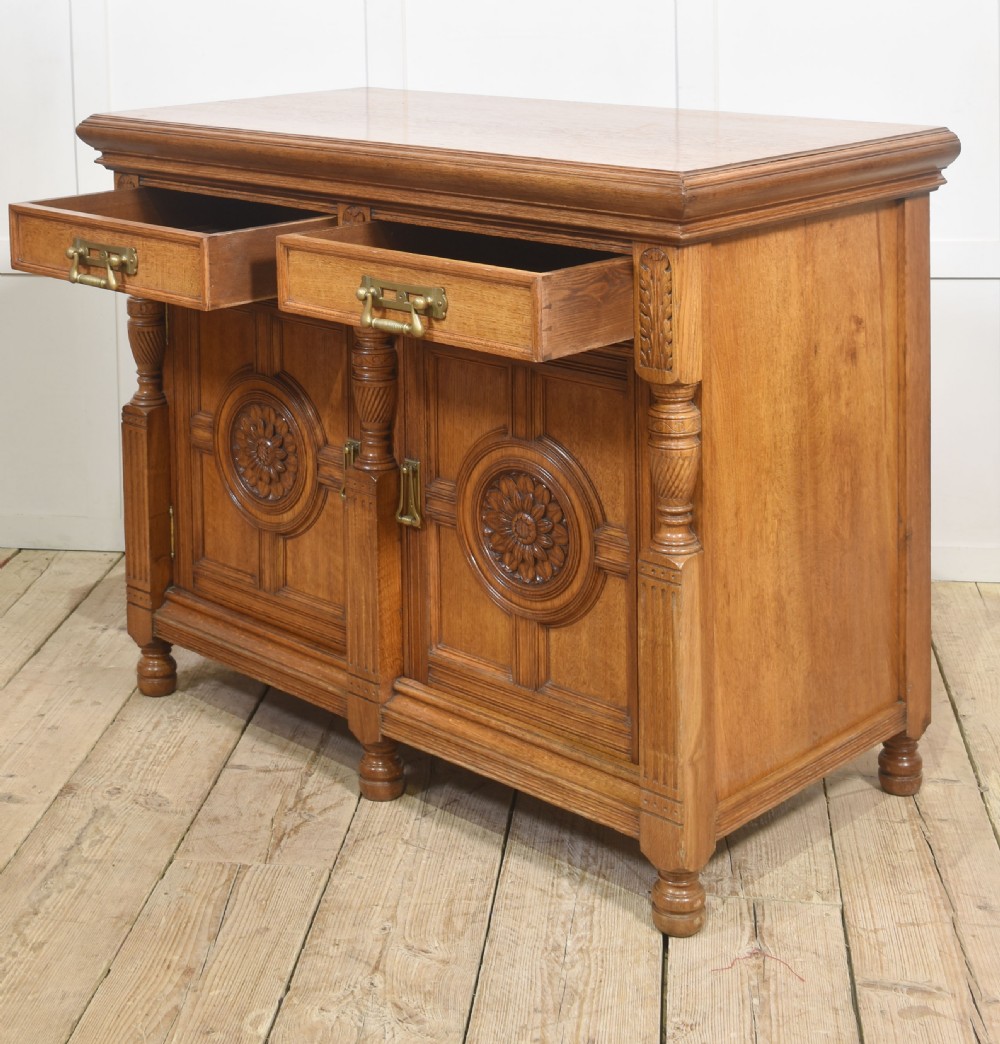 A carved oak two-door sideboard featuring two drawers with brass handles, central rosette carvings on the cupboard doors, and turned columns. View XVIII