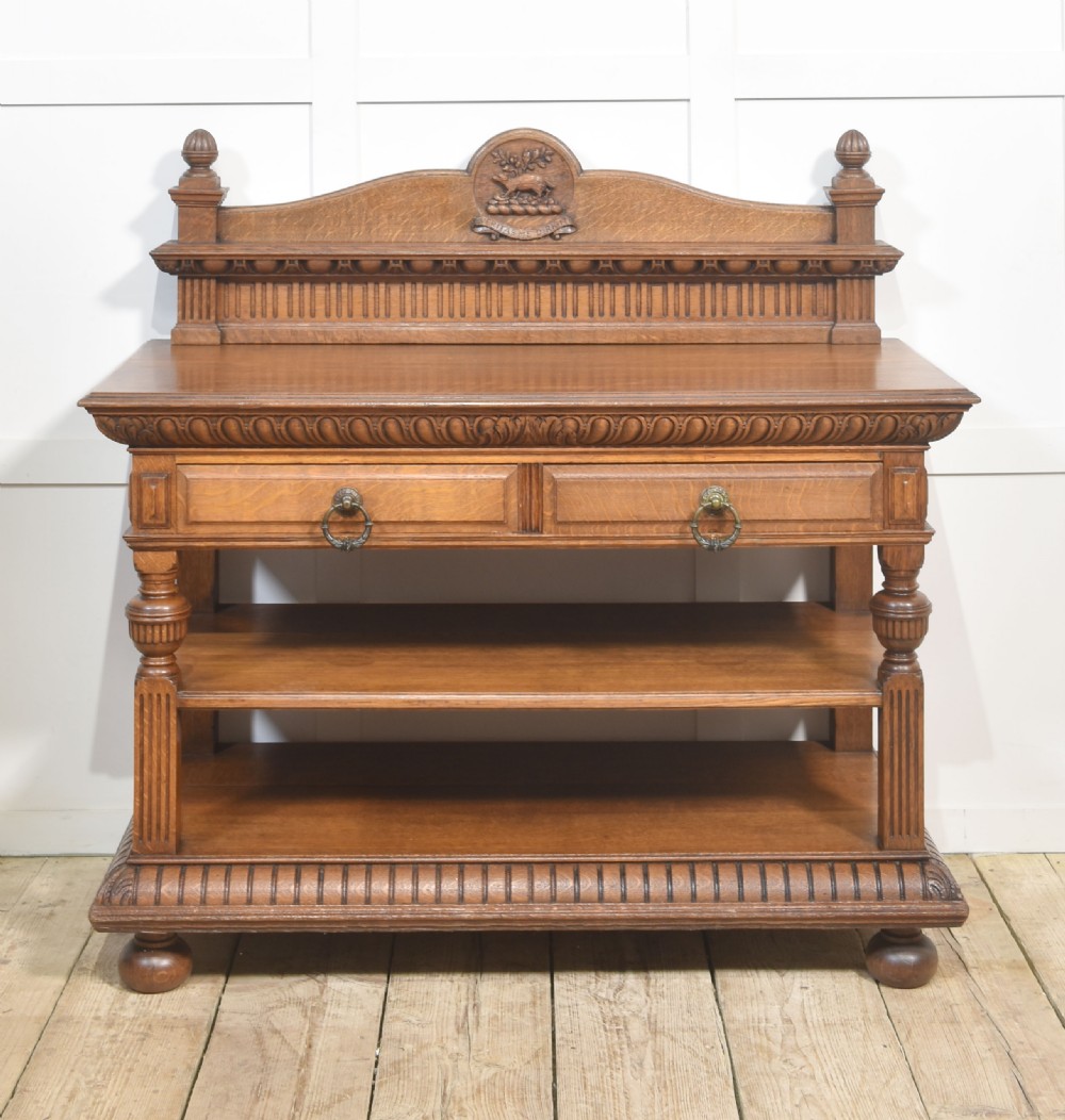 A carved oak serving table featuring a raised gallery back with a crest, two drawers with brass ring handles, and two open lower shelves supported by fluted turned legs.