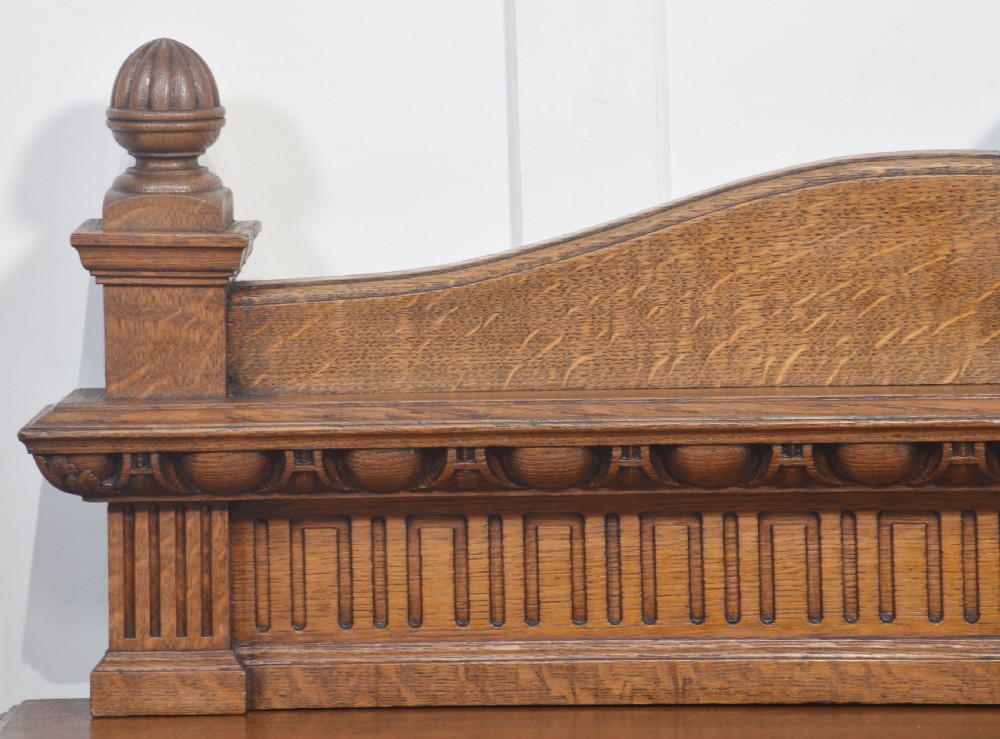 A carved oak serving table featuring a raised gallery back with a crest, two drawers with brass ring handles, and two open lower shelves supported by fluted turned legs. View II