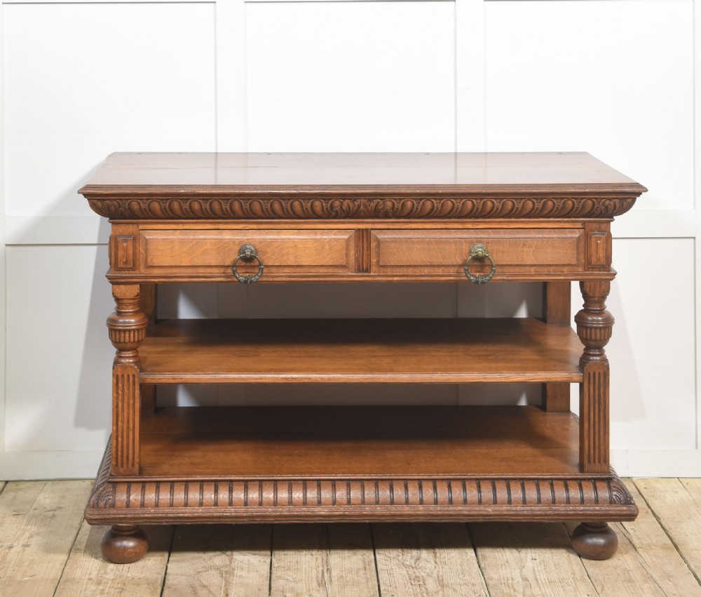 A carved oak serving table featuring a raised gallery back with a crest, two drawers with brass ring handles, and two open lower shelves supported by fluted turned legs. View V