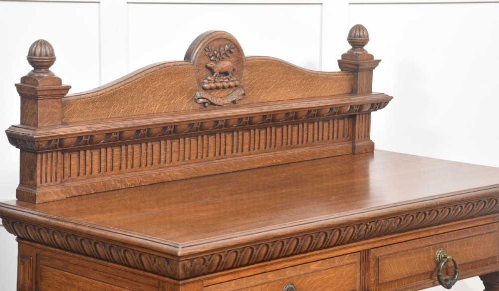 A carved oak serving table featuring a raised gallery back with a crest, two drawers with brass ring handles, and two open lower shelves supported by fluted turned legs. View VI