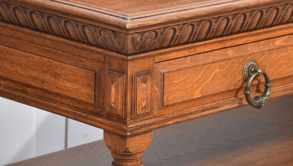 A carved oak serving table featuring a raised gallery back with a crest, two drawers with brass ring handles, and two open lower shelves supported by fluted turned legs. View VIII