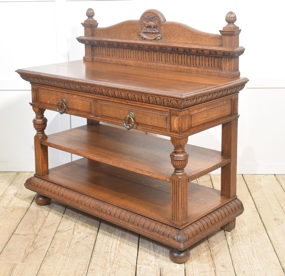 A carved oak serving table featuring a raised gallery back with a crest, two drawers with brass ring handles, and two open lower shelves supported by fluted turned legs. View X