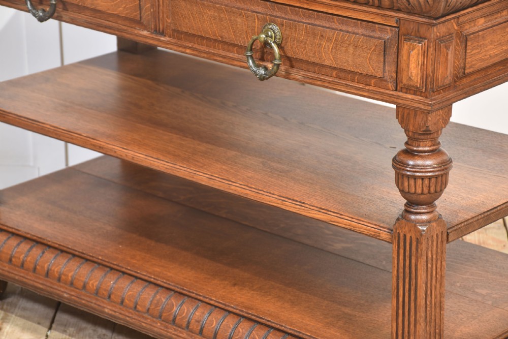 A carved oak serving table featuring a raised gallery back with a crest, two drawers with brass ring handles, and two open lower shelves supported by fluted turned legs. View XI