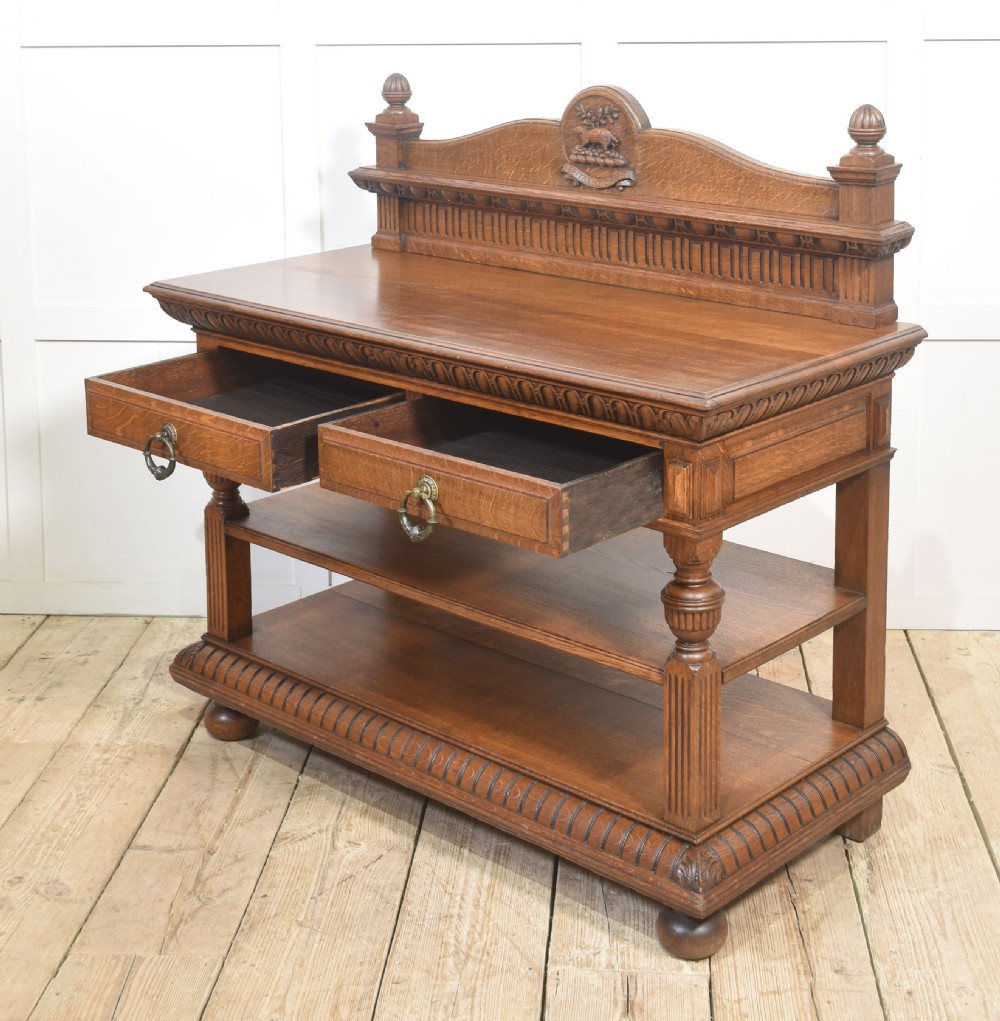A carved oak serving table featuring a raised gallery back with a crest, two drawers with brass ring handles, and two open lower shelves supported by fluted turned legs. View XIII