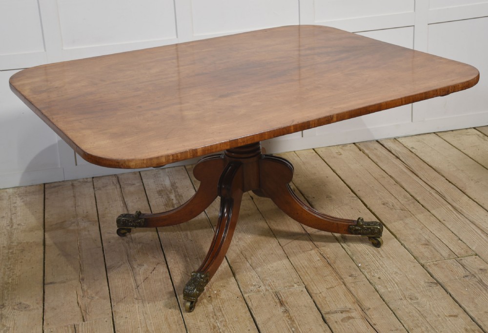 A Regency mahogany tilt-top breakfast table featuring a rectangular top with rounded corners, supported by a turned central pedestal and four splayed legs with brass paw castors.