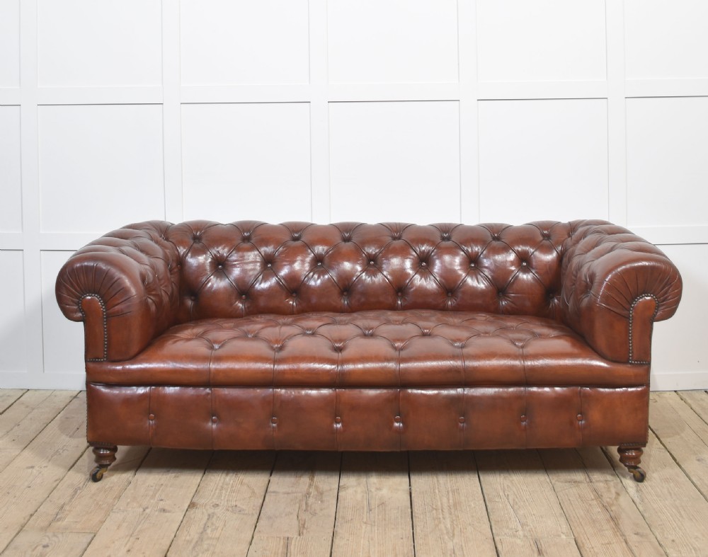 A chestnut brown leather Chesterfield sofa featuring deep buttoned upholstery, rolled arms, brass studding, and turned wood legs with castors.