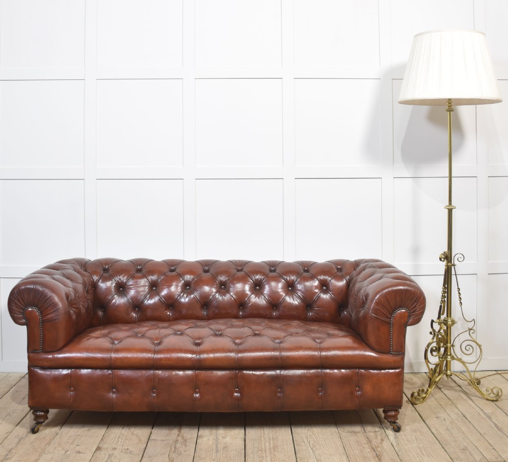 A chestnut brown leather Chesterfield sofa featuring deep buttoned upholstery, rolled arms, brass studding, and turned wood legs with castors. View II