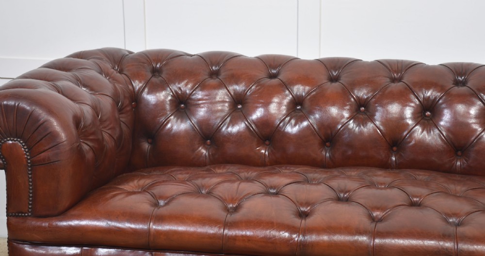 A chestnut brown leather Chesterfield sofa featuring deep buttoned upholstery, rolled arms, brass studding, and turned wood legs with castors. View III