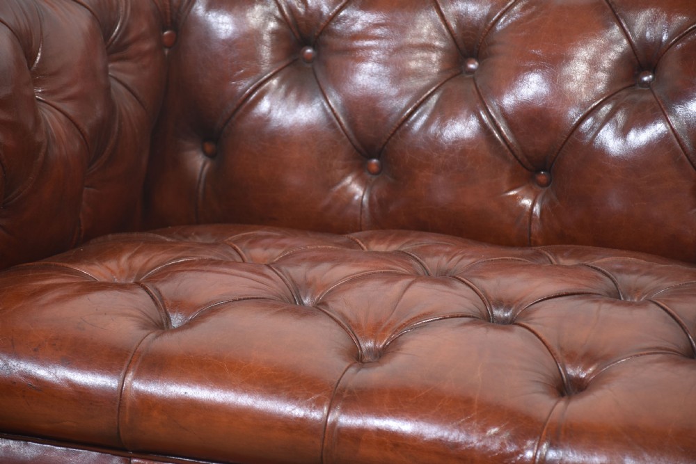 A chestnut brown leather Chesterfield sofa featuring deep buttoned upholstery, rolled arms, brass studding, and turned wood legs with castors. View V