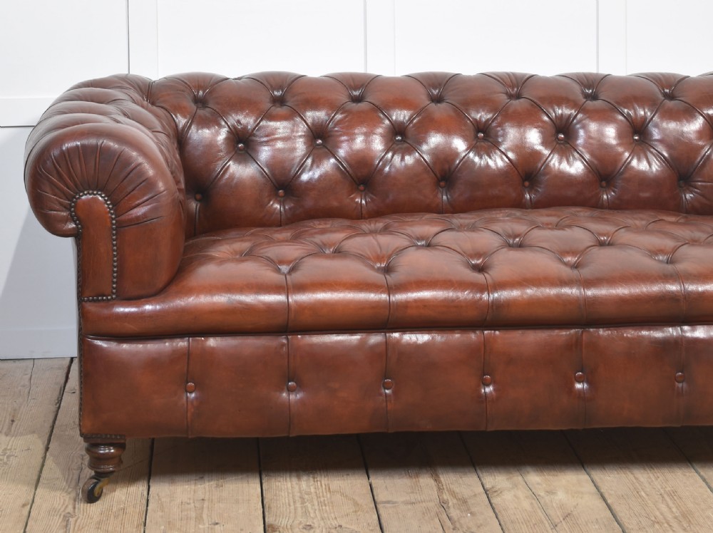 A chestnut brown leather Chesterfield sofa featuring deep buttoned upholstery, rolled arms, brass studding, and turned wood legs with castors. View VI
