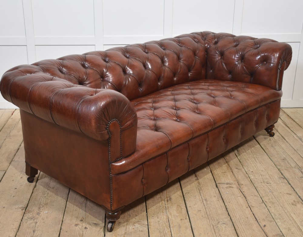 A chestnut brown leather Chesterfield sofa featuring deep buttoned upholstery, rolled arms, brass studding, and turned wood legs with castors. View VII