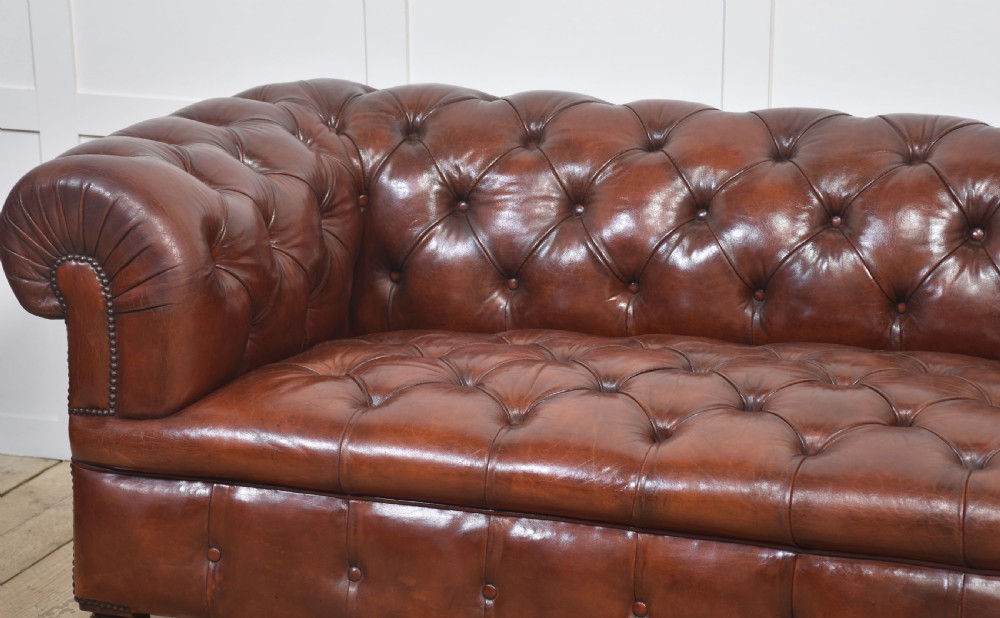A chestnut brown leather Chesterfield sofa featuring deep buttoned upholstery, rolled arms, brass studding, and turned wood legs with castors. View VIII