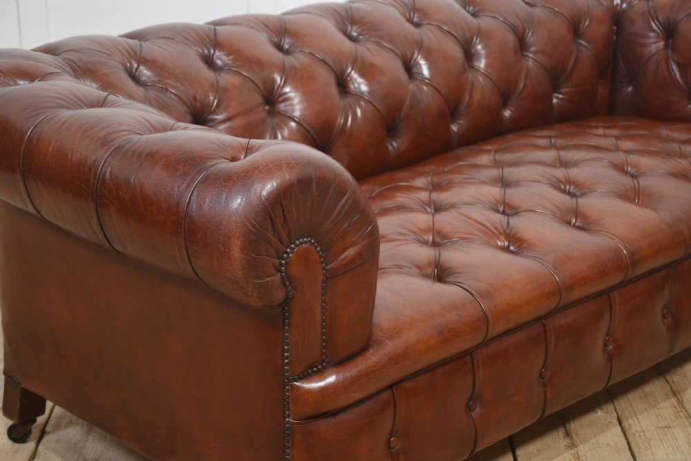 A chestnut brown leather Chesterfield sofa featuring deep buttoned upholstery, rolled arms, brass studding, and turned wood legs with castors. View IX