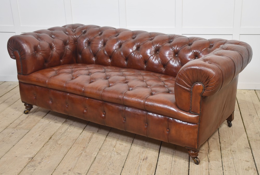 A chestnut brown leather Chesterfield sofa featuring deep buttoned upholstery, rolled arms, brass studding, and turned wood legs with castors. View X