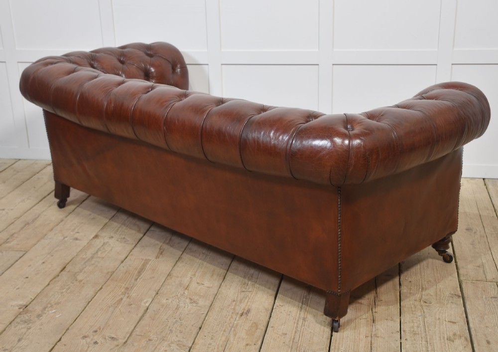 A chestnut brown leather Chesterfield sofa featuring deep buttoned upholstery, rolled arms, brass studding, and turned wood legs with castors. View XI