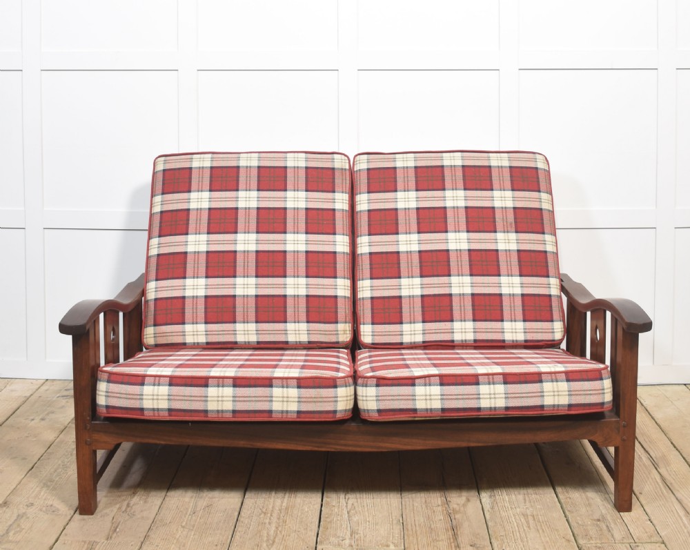 An Arts and Crafts solid walnut two-seater settee with red and cream tartan plaid upholstered cushions.
