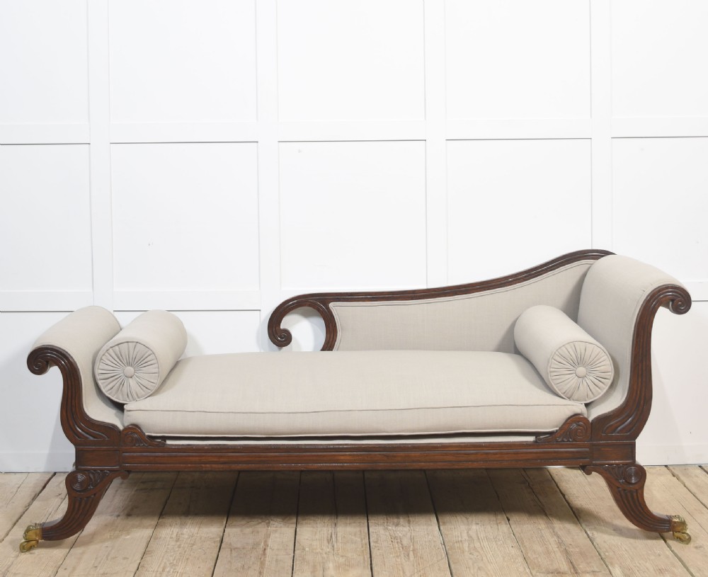 A Regency rosewood chaise longue with carved scroll arms, light grey fabric upholstery, matching bolster cushions, and brass castors.