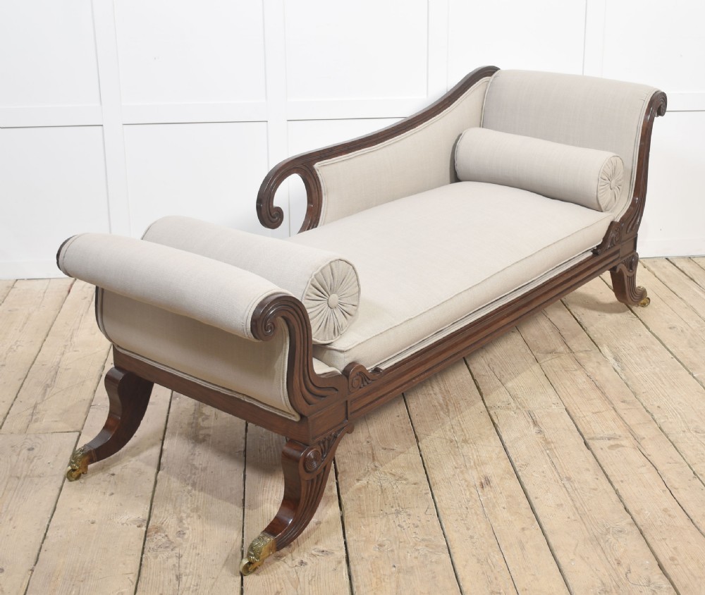 A Regency rosewood chaise longue with carved scroll arms, light grey fabric upholstery, matching bolster cushions, and brass castors. View III