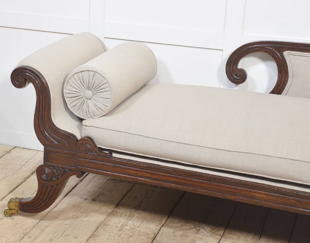 A Regency rosewood chaise longue with carved scroll arms, light grey fabric upholstery, matching bolster cushions, and brass castors. View V