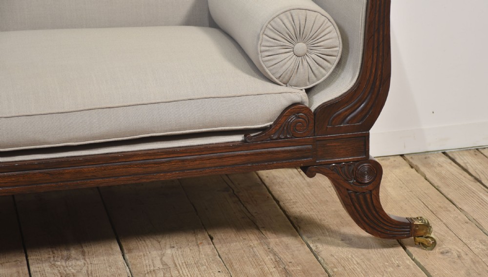 A Regency rosewood chaise longue with carved scroll arms, light grey fabric upholstery, matching bolster cushions, and brass castors. View VI