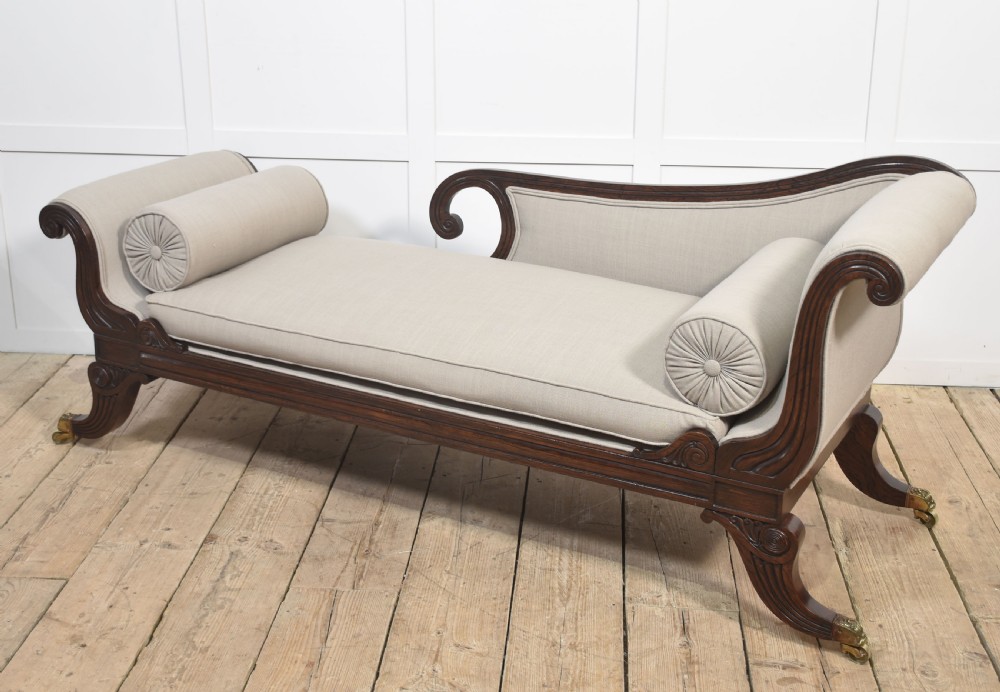 A Regency rosewood chaise longue with carved scroll arms, light grey fabric upholstery, matching bolster cushions, and brass castors. View X