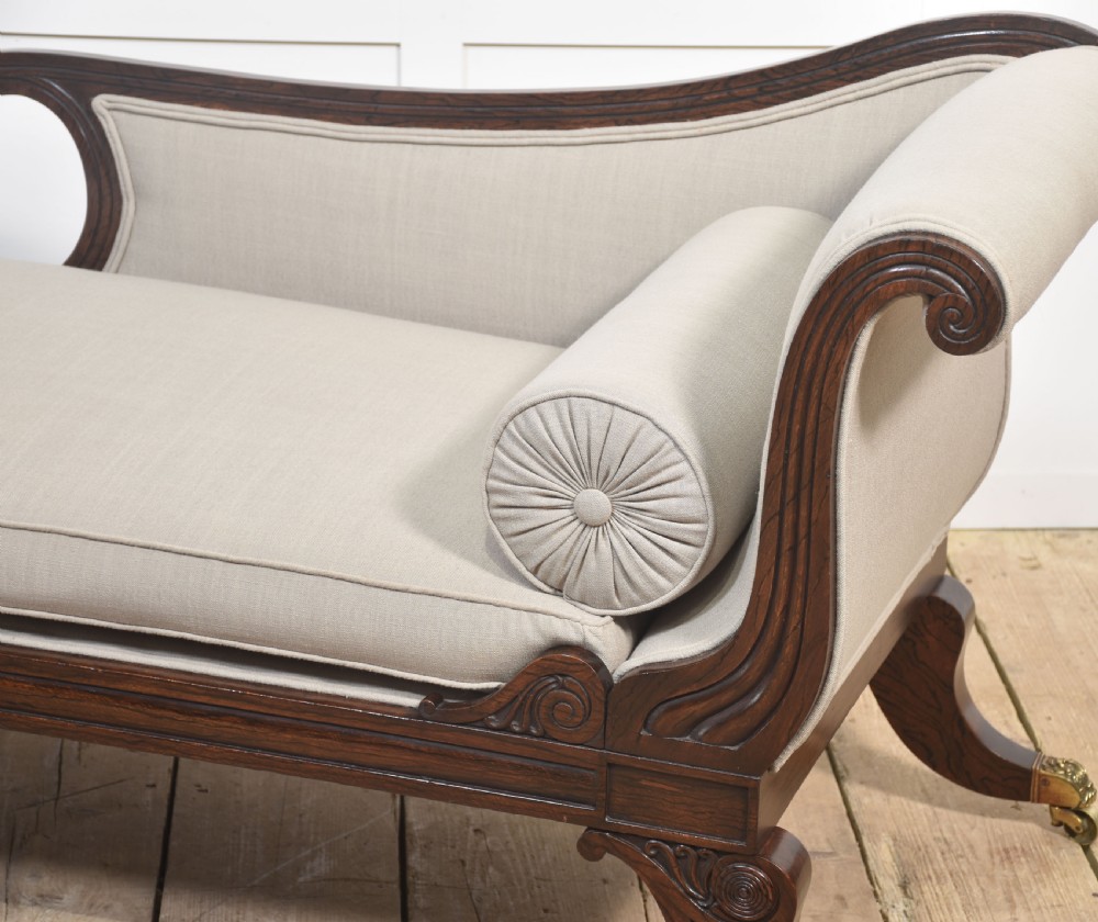 A Regency rosewood chaise longue with carved scroll arms, light grey fabric upholstery, matching bolster cushions, and brass castors. View XI