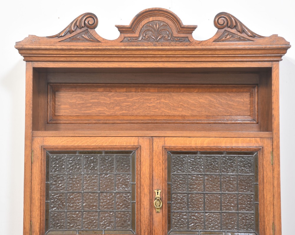 A warm brown oak dresser featuring a carved scrollwork pediment, leaded glass upper cabinet doors, a central mirror insert, a single long drawer, and double lower doors with geometric panel carving. View III